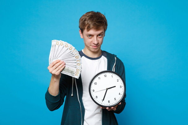 Confident young man in casual clothes holding fan of money in dollar banknotes, cash money, round clock isolated on blue wall background. People sincere emotions lifestyle concept. Mock up copy space - Fotó, kép