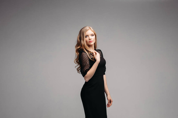 Attractive serious woman in long black dress posing in studio and looking at camera. Gorgeous model with beautiful makeup and hairstyle standing in front of grey isolated background. - Zdjęcie, obraz