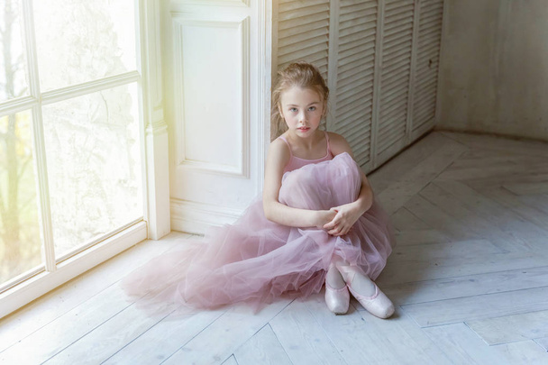 Young classical ballet dancer girl in dance class. Beautiful graceful ballerina in pink tutu skirt puts on pointe shoes near large window in white light hall - Photo, Image