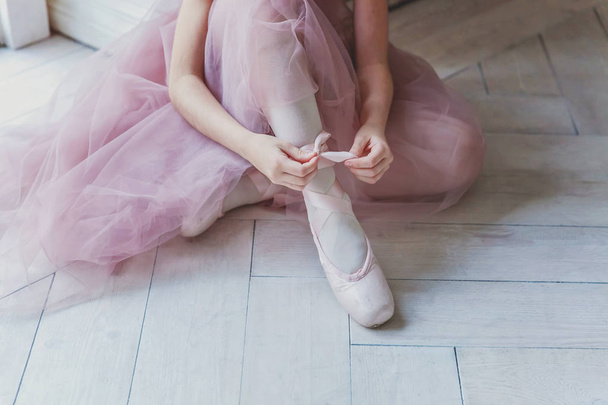 Руки балерины в розовой юбке в балетной пачке надевают пуанты на ноги в белом светлом зале. Молодая танцовщица классического балета - Фото, изображение