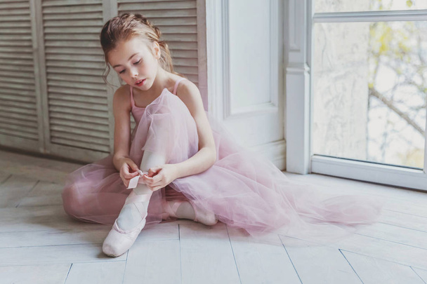 Молодая танцовщица балета в танцевальном классе. Красивая изящная балерина в розовой юбке в пачке надевает пуанты рядом с большим окном в белом светлом зале - Фото, изображение