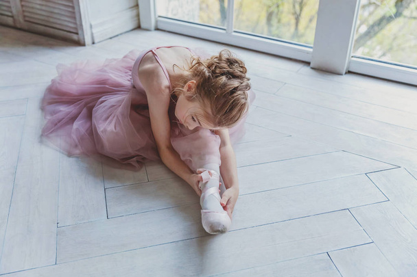 Молодая танцовщица балета в танцевальном классе. Прекрасная изящная балерина практикует балетные позиции в розовой юбке в белом светлом зале
 - Фото, изображение