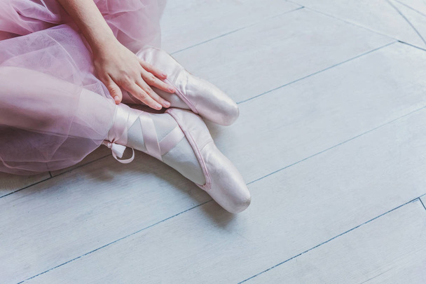 Hands of ballerina in pink tutu skirt puts on pointe shoes on leg in white light hall. Young classical ballet dancer girl in dance class - Foto, Imagen
