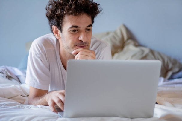 Happy young man using laptop and relaxing on his bed. Indoors - Foto, Imagen