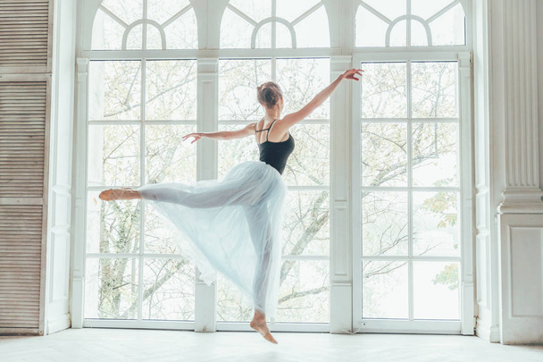 junge klassische Balletttänzerin im Tanzkurs. schöne anmutige Ballerina übt Ballettpositionen in blauem Tutu-Rock in der Nähe eines großen Fensters im weißen Lichtraum - Foto, Bild