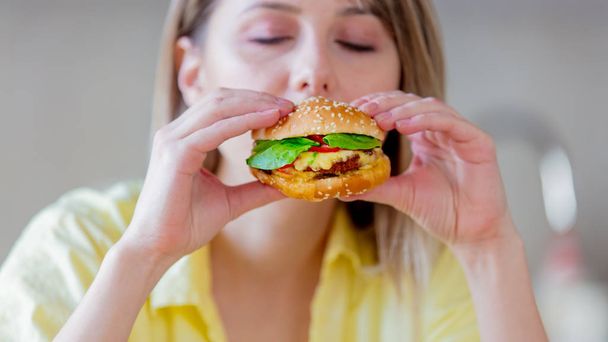 Young beautiful woman eating handmade hamburger at kitchen  - Фото, изображение
