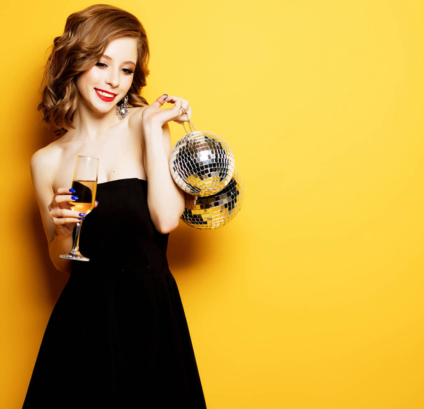 sexy woman with a glass of white wine and disco ball over yellow background - Valokuva, kuva