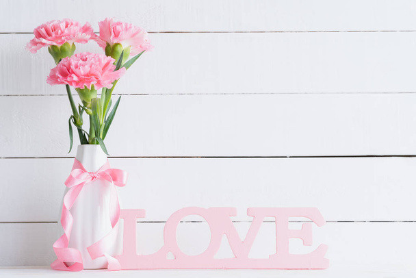 Valentines day and love concept. Pink carnation flower in vase with Wooden letters forming word LOVE written on white background. - Fotó, kép