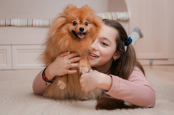 Teenage girl with a dog breed Spitz rejoices with a pet at home on the floor. Care and training of a pet. Content Fluffy puppy. To groom long-haired Pomeranian. Orange red - Photo, Image