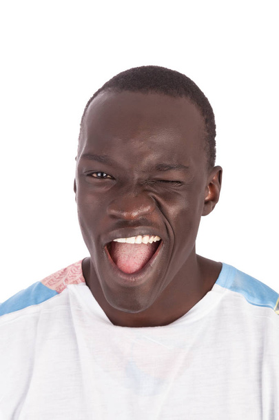 Joven hombre sudanés guapo cara a cámara en camiseta blanca guiñando el ojo, sonriendo con dientes blancos y lengua rosa mostrando, boca de cara recta abierta en formato retrato con espacio de copia aislado en blanco
. - Foto, Imagen