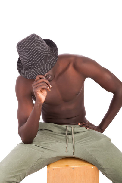 Joven aficionado hombre sudanés con un torso desnudo en pantalones caqui usando un sombrero casualmente sentado en un tambor de cajón en una postura abierta de bienvenida. La imagen está en formato vertical con espacio de copia y sujeto aislado en blanco. Inspiración para meme
. - Foto, imagen