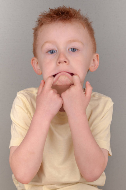 Fiery redhead young boy fingers in his mouth pulling lips down in sad face concept wearing a yellow shirt in portrait format with copy space. Meme or poster of cute redhead cheeky sad, bad tempered. - Photo, Image