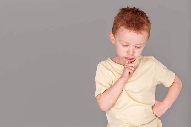 Fiery redhead young boy in yellow shirt in finger to chin in deep thought stance in landscape format on grey background with copy space. Metaphor for thinking. Meme and poster image for cute redhead. - Foto, Imagen