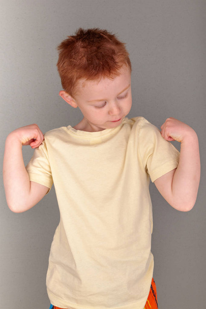 Fiery redhead young boy in yellow shirt looking at left arm with strong arms pose, portrait format on grey background with copy space. Being strong and coping with all of life's adventures metaphor. - Fotó, kép