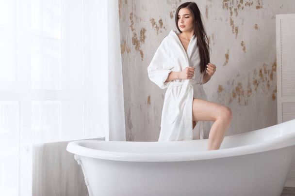Young woman in a white bathrobe sitting at the side of freestanding white bath - Fotoğraf, Görsel