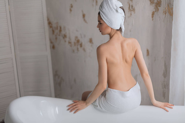 Young woman with naked back wearing towel on head, sitting on bathtub edge. - Foto, Imagem