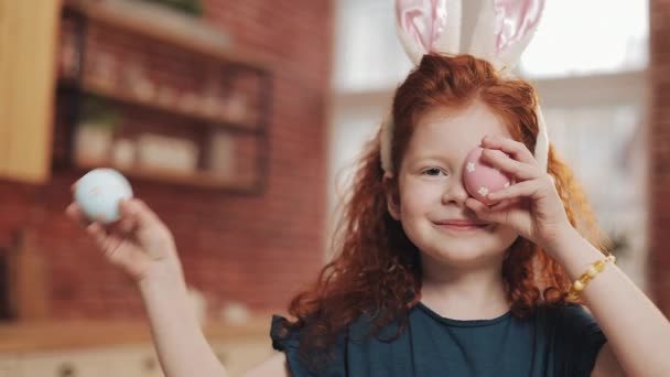 Portrait of cheerful redhead little kid girl with bunny ears with an easter egg on the kitchen background. She is cheering and having fun at the camera. Happy Easter - Materiaali, video