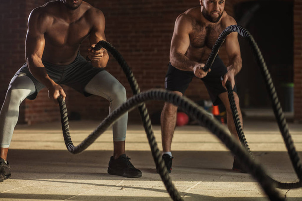 Two multiethnic bare chest sportsmen exercising with battle ropes at the gym. - Foto, Imagen