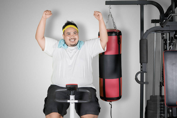 Picture of young obese man expressing happiness while using an exercise bike in the studio - Valokuva, kuva