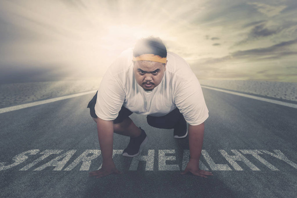 Picture of an obese man ready to run while kneeling above text of start healthy on the road. Shot with sunlight background - Photo, Image