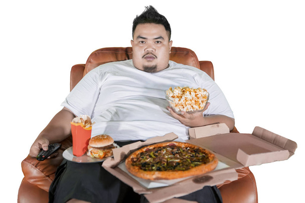 Overweight man eating junk foods while watching TV and sitting on the sofa, isolated on white background - Photo, image