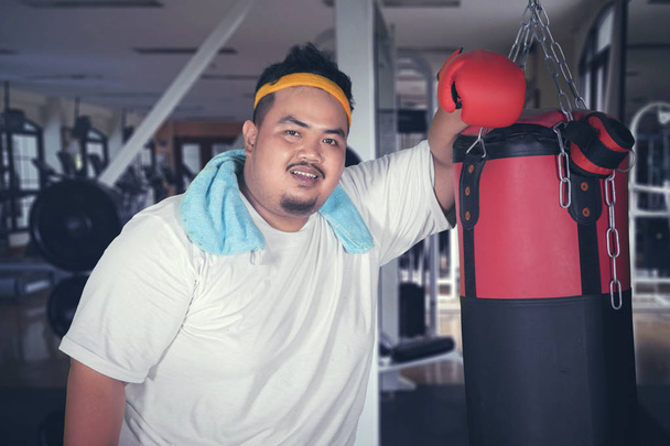 Picture of overweight man smiling at the camera while leaning on a boxing bag. Shot in the gym center - Φωτογραφία, εικόνα