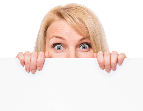 Young woman with surprised eyes peeking out from behind billboard paper poster. Businesswoman holding big white banner, isolated on white background. - Photo, image