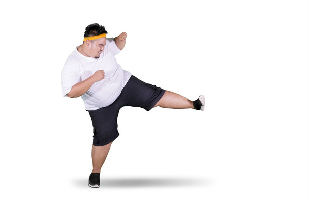 Portrait of a young fat man wearing sportswear while performing a kick in the studio, isolated on white background - Фото, изображение