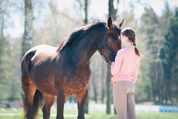 Young teenage girl standing with her favorite brown horse. Colored outdoors horizontal springtime image with filter - Photo, Image