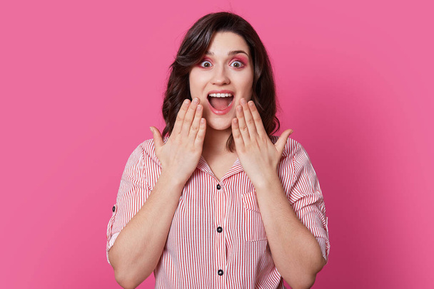 Photo of attractive overemotive young lady with dark hair, opens mouth widdely, keeps hands on cheeks, has pink makeup, reacts on rumors, wears stylish shirt, models indoor. Emotions concept - Photo, image