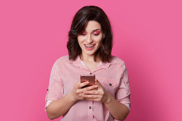 Technologically educated female person is in center of portrait photography, holds in hands, clasps unknown brand smartphone with her fingers while she is pleasantly surprised at this situation. - Photo, image