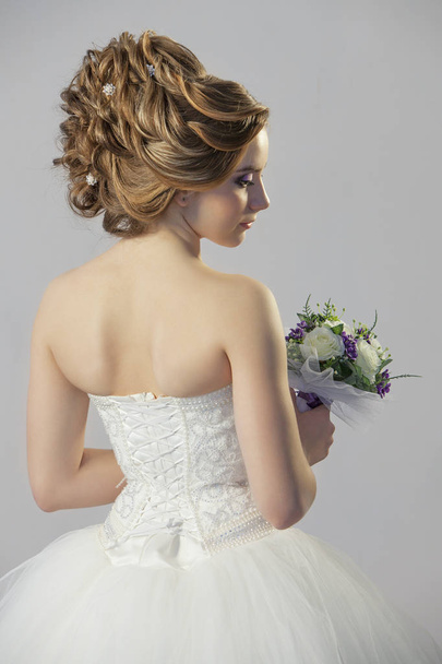 young bride girl in white dress with a bouquet of flowers shows a beautiful wedding hairstyle on a gray background - Zdjęcie, obraz