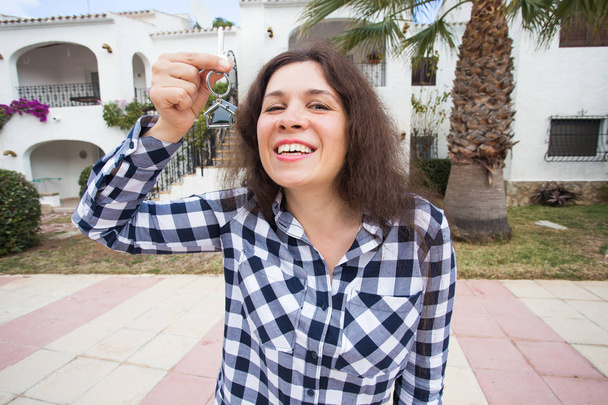 New home, house, property and tenant - Young funny woman holding key in front of her new home after buying real estate - Photo, Image