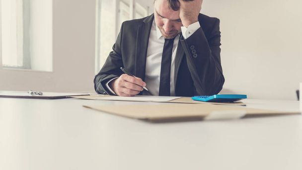 Übermüdeter Geschäftsmann, der an seinem Schreibtisch sitzt und seinen Kopf mit dem Arm stützt, während er versucht, ein Problem zu lösen, indem er Papierkram und Bericht durchsieht. - Foto, Bild