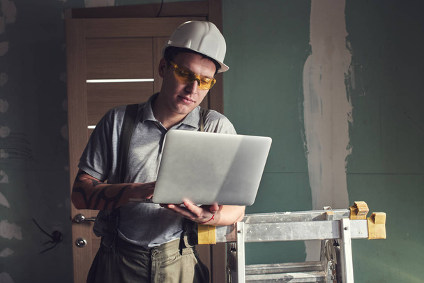 Room repair. Builder in helmet and glasses stands with a laptop in his hands on the background of the construction - Foto, Imagem
