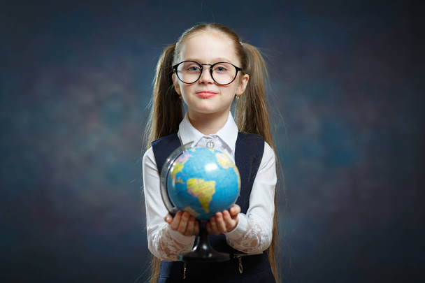 Блондинка-школьница держит в руках глобус мира. Молодая женщина начинает изучать географию. Красотка с двумя длинными хвостиками в школьной форме Посмотрите на модель карты Земли
 - Фото, изображение