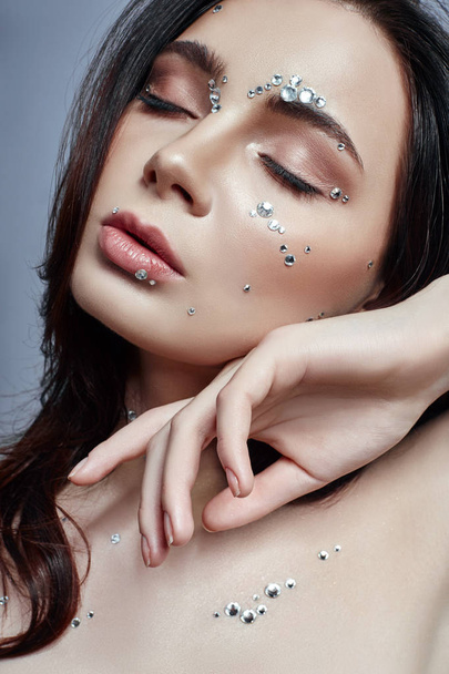 Art portrait of a woman with large crystals of rhinestones on her face, chest and body. Perfect makeup, skin care, professional cosmetics. Girl's face with diamonds on the skin close-up - Φωτογραφία, εικόνα
