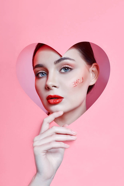 Woman looking in the heart hole, bright beautiful makeup, big eyes and lips, bright lipstick, professional cosmetics and facial care. Bright colored background and a gap slot in the paper heart - Fotó, kép