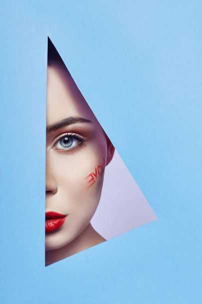 Woman looking in blue triangle hole, bright beautiful makeup love word, big eyes and lips, bright lipstick, professional cosmetics and facial care. Bright colored background gap slot in paper triangle - Photo, Image
