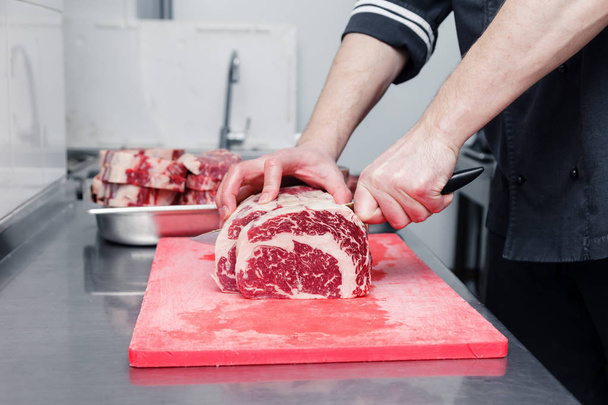 Szekrény szakács kézzel vágja bordás márvány marhahús steak éles kés piros műanyag vágódeszka fém asztalon étterem konyhájában. Koncepció steakhouse specializálódott grillezett hús, élő tűz - Fotó, kép