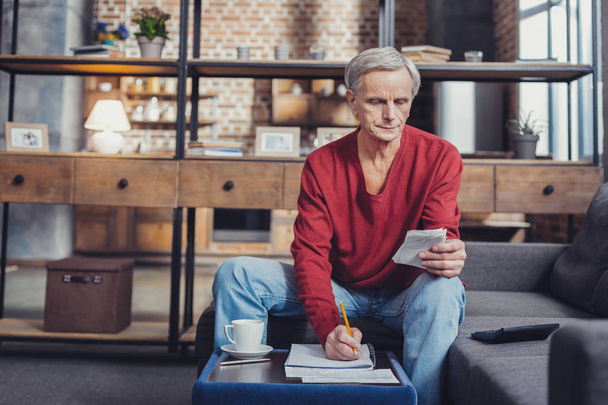 Senior man making notes while sitting in his living room - Photo, Image