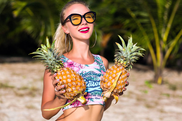 Inspired european girl in trendy sunglasses holding pineapples while posing at beach. Joyful white girl in stylish tank-top having fun during summer vacation and enjoying fruits. - Foto, imagen