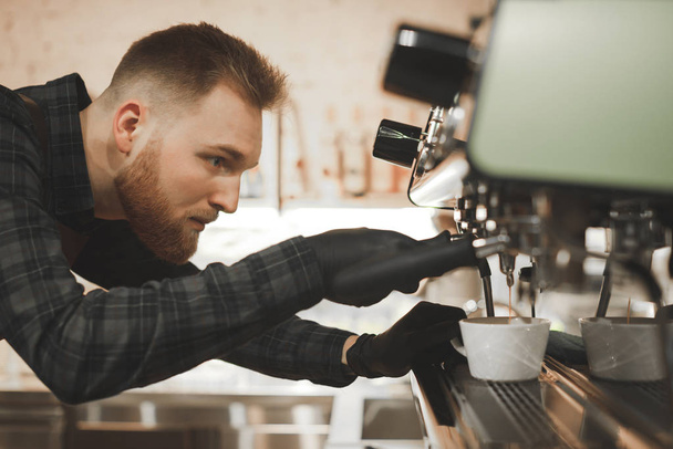 Bir kafede kapuçino hazırlanıyor sakallı konsantre barista portresi kapatın. Cafe sahibi bir kahve makinesi kahve yapmaya odaklanmış, kahve bir fincan içine dökülür gibi görünüyor - Fotoğraf, Görsel
