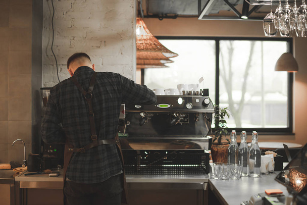 Professional barista preparing coffee in the coffee machine in a cozy cafe. View from the back. Coffee culture, professional making, service and catering concepts. Copyspace - Foto, Imagen