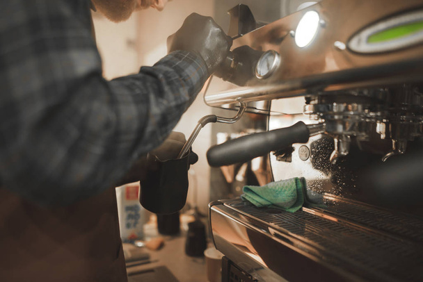 Close-up photo, barista steaming milk in pitcher on a professional coffee machine. Barista professional cooks coffee in the coffee shop. Man makes a cappuccino on a professional coffee machine. - Photo, Image
