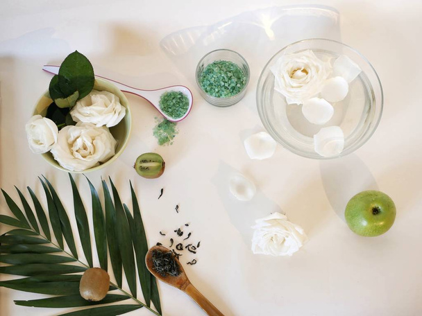 Weiße Rosen in Wasser, Blütenblätter, grüne Früchte, Meersalz, grüner Tee auf einem hellen Tisch, Draufsicht, Maskenzutaten, sanfte Pflegeverfahren - Foto, Bild