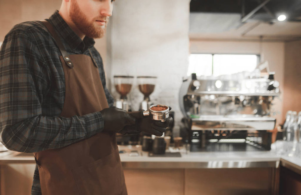 Baristan parrakas mies kahvilassa ja pitää käsissään portafiltteriä, jossa on jauhettua kahvia. Kahvilan omistaja valmistaa kahvia. Kopioi
 - Valokuva, kuva