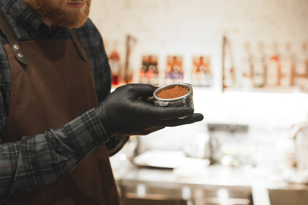 Foto ravvicinata di un barista con la barba che tiene un portafiltro e prepara il caffè nella sua caffetteria. Barista tiene un supporto con caffè macinato
. - Foto, immagini