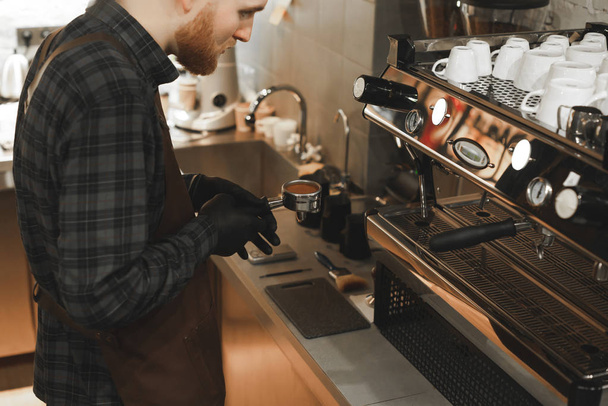 Nahaufnahme eines bärtigen Baristas, der einen Filter an der Kaffeemaschine im Café hält. Baristas Hände halten einen Halter mit geröstetem Kaffee im Café. Kaffee-Konzept. - Foto, Bild