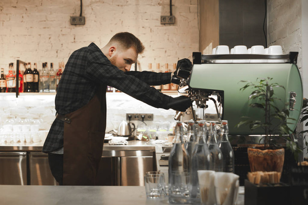 Retrato de barista barbudo concentrado preparando capuchino en una cafetería. Propietario del café se centra en hacer café en una máquina de café
. - Foto, Imagen